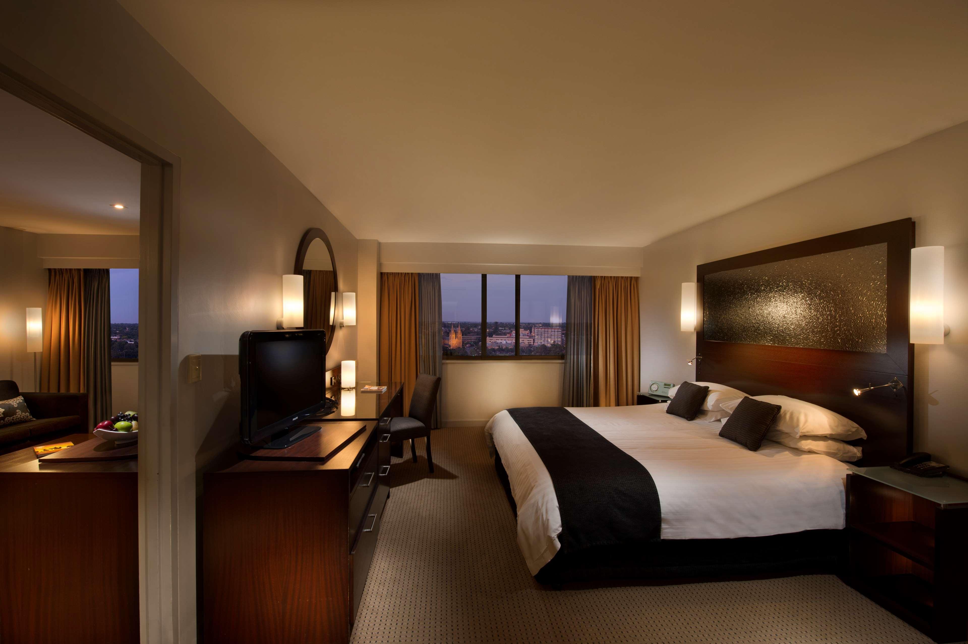Stamford Plaza Adelaide Hotel Kültér fotó The photo features a modern hotel room that includes a large bed with dark and light bedding, complemented by two pillows. The room is well-lit with warm lighting and has a contemporary aesthetic. To one side, there is a wooden dresser with a flat-sc