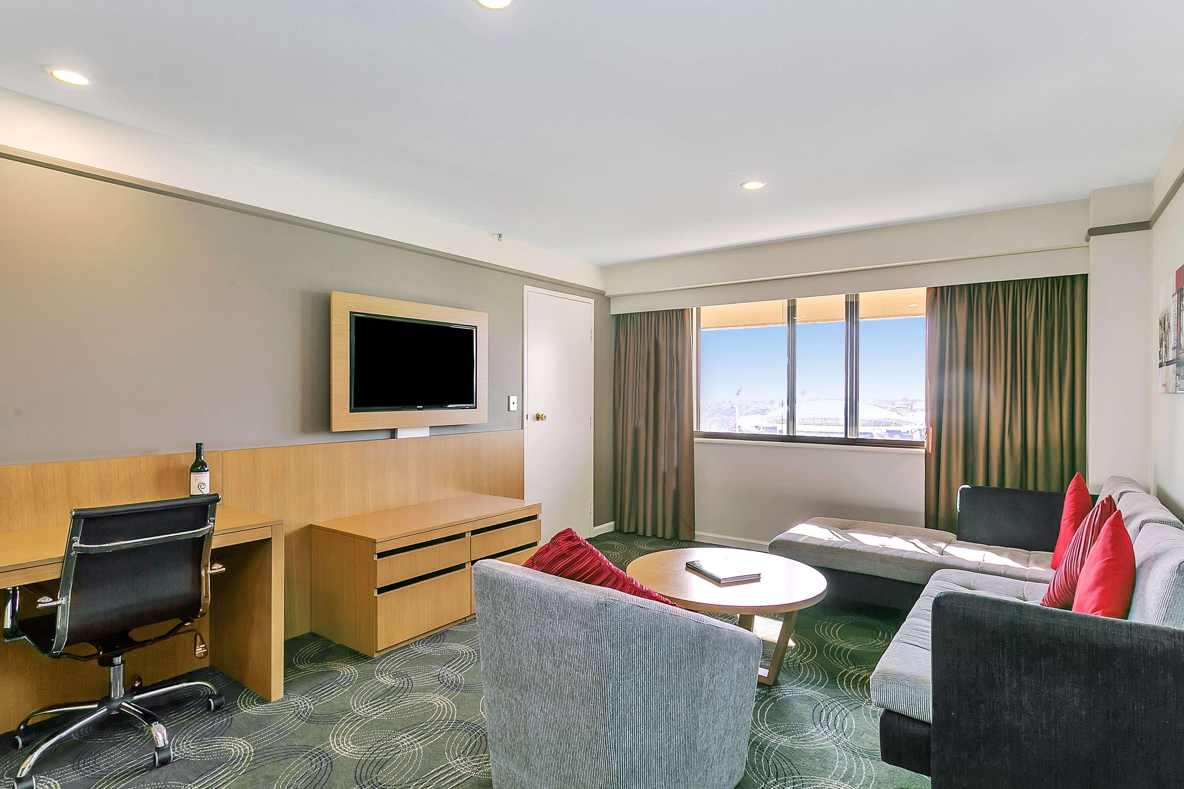 Stamford Plaza Adelaide Hotel Kültér fotó The photo shows a modern hotel room or suite that features a cozy seating area. There is a comfortable sofa with red accent pillows, a round coffee table in the center, and a sleek desk with an office chair. On the wall, there is a flat-screen TV mou