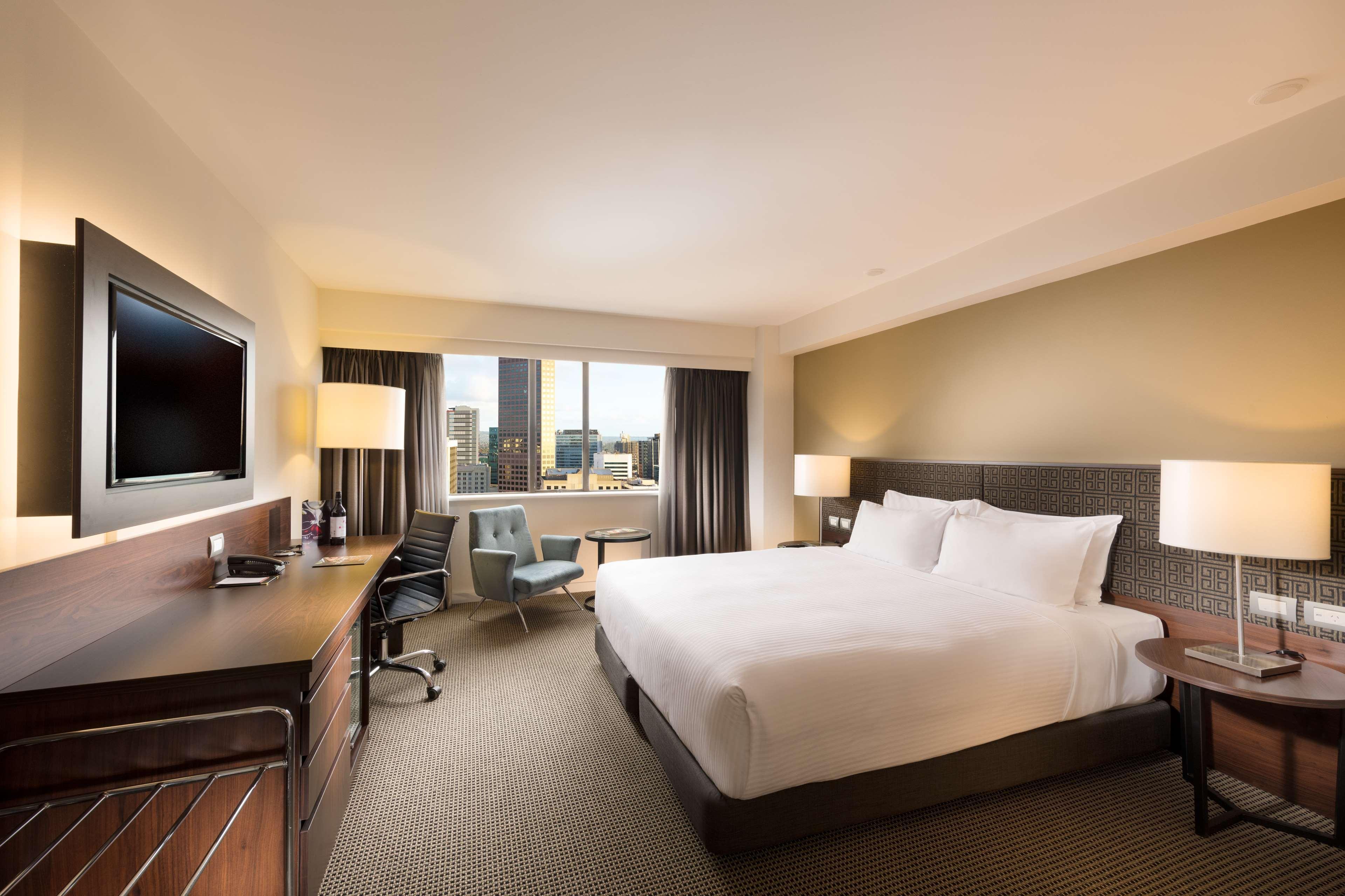 Stamford Plaza Adelaide Hotel Kültér fotó The photo shows a modern hotel room with a contemporary design. It features a large bed with white linens, a bedside table on either side with lamps, and a dark wood accent wall. There's a work desk with an office chair and a flat-screen television m