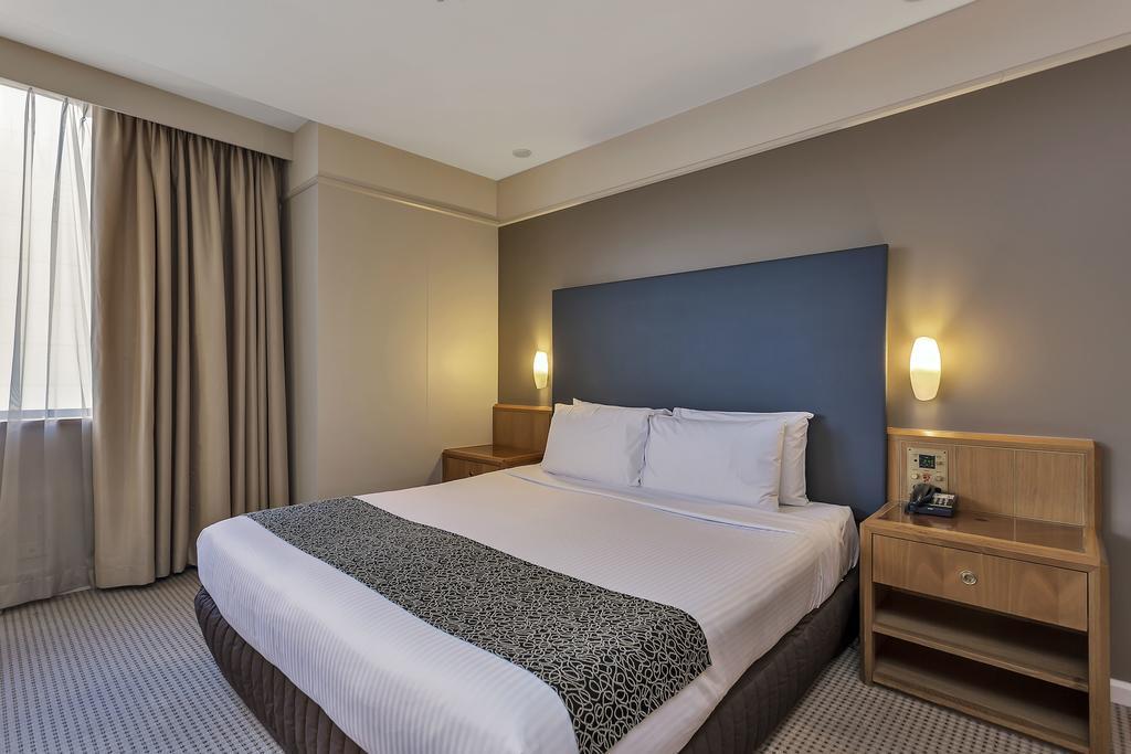 Stamford Plaza Adelaide Hotel Kültér fotó The photo shows a modern hotel room. It features a double bed with a white duvet and a decorative grey and black throw. The headboard is a solid dark color. There are two bedside tables on either side of the bed, each with a lamp. Natural light enter